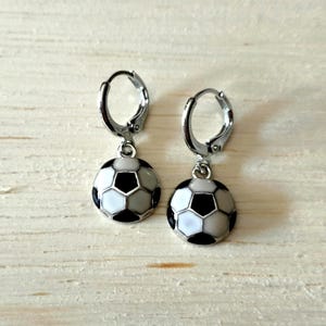 Puede incluir: Pendientes de aro plateados con colgantes de balón de fútbol. Cada colgante es un pequeño balón de fútbol con pentágonos y hexágonos negros y blancos. Se muestran sobre una superficie de madera clara.