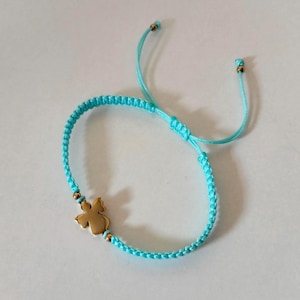 Puede incluir: Una pulsera de macram&eacute; turquesa con un dije dorado en forma de flor. La pulsera tiene nudos corredizos ajustables y peque&ntilde;as cuentas doradas. El fondo es una superficie blanca lisa.