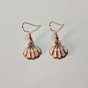 Könnte beinhalten: Ein Paar Muschelohrringe. Jeder Ohrring hat einen goldfarbenen Haken und einen muschelförmigen Anhänger. Die Anhänger sind weiß mit einem rosa Zentrum und goldfarbenen Verzierungen. Die Ohrringe sind für durchbohrte Ohren.