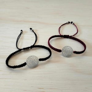 Könnte beinhalten: Zwei verstellbare Armbänder mit geflochtenem Kordeldesign. Ein Armband ist schwarz, das andere weinrot. Jedes Armband hat einen silberfarbenen runden Anhänger mit einem Spiralmuster. Die Armbänder werden auf einer hellen Holzoberfläche präsentiert.