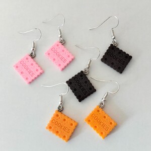 Puede incluir: Un conjunto de pendientes colgantes con forma de galletas cuadradas. Los pendientes son de color rosa, negro y naranja, cada uno con la palabra "COOKIES" impresa. Tienen ganchos plateados.