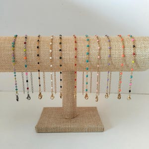 Peut inclure: Une collection de onze bracelets à chaîne délicats présentés sur un support en T. Chaque bracelet comporte de petites perles colorées espacées le long d'une fine chaîne métallique, incluant des tons turquoise, rose, noir, blanc, rouge, orange et violet.