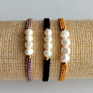 Op de afbeelding: Drie handgemaakte armbanden met witte parels en gouden accenten. De armbanden zijn in de kleuren mauve, zwart en goud. De armbanden worden gepresenteerd op een beige, getextureerd oppervlak.