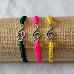 May include: Three friendship bracelets with a silver treble clef charm. The bracelets are green, pink, and yellow.