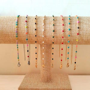 Op de afbeelding: Een display van kleurrijke kettingen met kralen. De kettingen hebben kralen in verschillende kleuren, waaronder turquoise, rood, zwart, wit en groen, geregen op goudkleurige kettingen. Elke ketting heeft een sluiting.