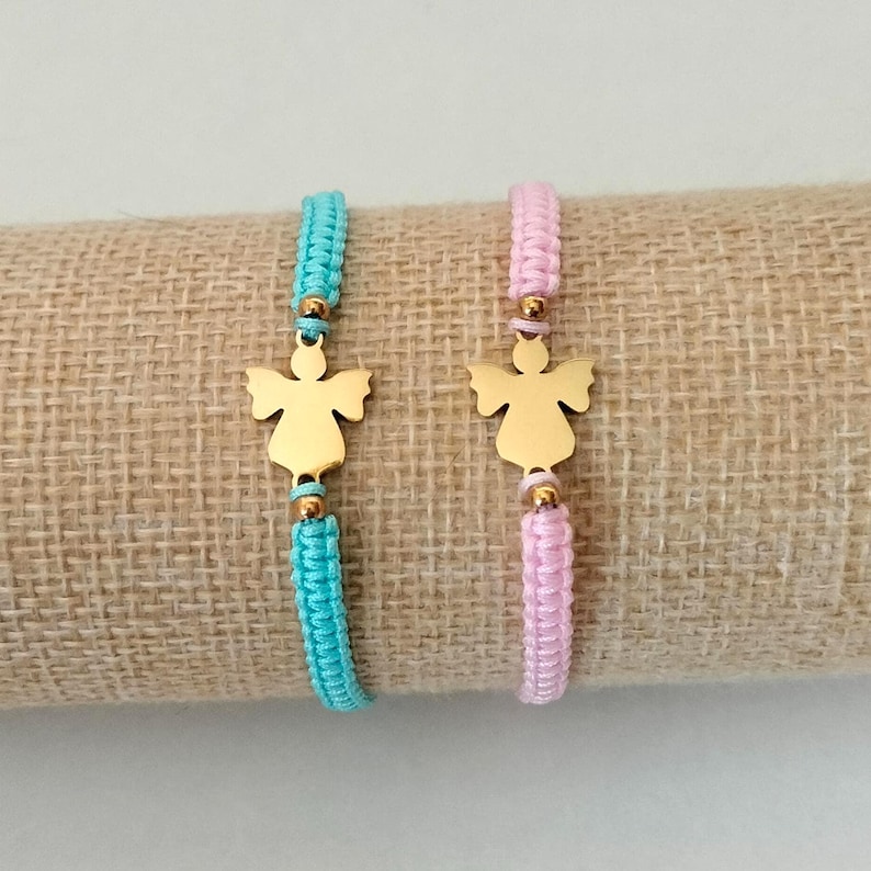 Puede incluir: Dos pulseras hechas a mano con colgantes de &aacute;ngel dorados. Una pulsera es azul claro, la otra rosa. Ambas pulseras est&aacute;n hechas con cord&oacute;n trenzado y cuentas doradas. Las pulseras se exhiben sobre una tela beige.
