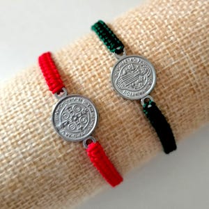 Könnte beinhalten: Zwei Armbänder mit silberfarbenen Medaillons und geflochtenen Kordeln. Eines hat eine rote Kordel, das andere eine grüne Kordel. Die Medaillons haben geprägte Designs und sind mit kleinen Metallschlaufen an den Kordeln befestigt. Der Hintergrund ist ein beiger Stoff.