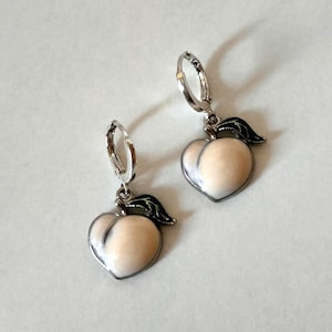 Puede incluir: Un par de pendientes con forma de melocotón con aros plateados. Los pendientes presentan un esmalte de color melocotón claro con un contorno plateado y un acento de hoja negra. Los pendientes están diseñados para colgar.