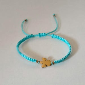 Puede incluir: Una pulsera de macram&eacute; turquesa con un dije dorado en forma de &aacute;ngel estilizado. La pulsera tiene nudos corredizos ajustables y peque&ntilde;as cuentas doradas. El fondo es una superficie blanca lisa.