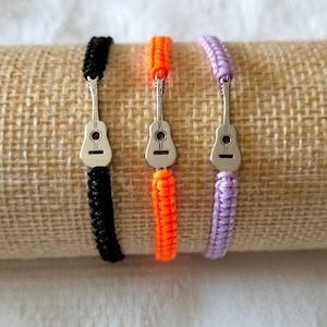 Può includere: Tre braccialetti dell'amicizia con ciondoli a forma di chitarra. I braccialetti sono neri, arancioni e viola.