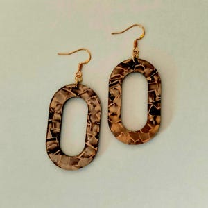 Puede incluir: Un par de pendientes ovalados con un diseño de mosaico en tonos marrones y beige. Los pendientes tienen ganchos dorados. El patrón de mosaico está formado por pequeñas formas irregulares, creando una apariencia texturizada. Aproximadamente 5 cm de largo.