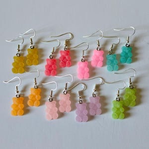 Puede incluir: Una colección de pendientes de gominolas de colores. Los pendientes presentan una variedad de colores, incluyendo amarillo, naranja, rosa, rojo, morado, verde y turquesa. Cada pendiente tiene un gancho plateado.