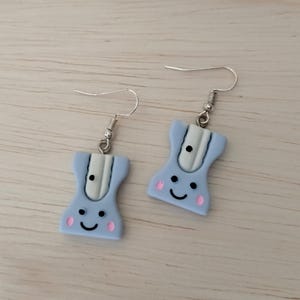 Könnte beinhalten: Ein Paar hellblaue Ohrringe in Form von Anspitzern. Jeder Ohrring hat eine weiße Mitte, schwarze Punkte, rosa Wangen und ein lächelndes Gesicht. Sie hängen an silberfarbenen Fischhaken-Ohrdrähten.