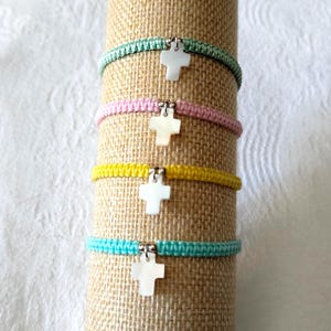 Könnte beinhalten: Vier Armbänder mit einem weißen Kreuzanhänger. Die Armbänder sind aus geflochtener Schnur in Grün, Rosa, Gelb und Blau.