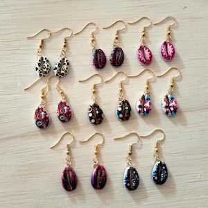 Può includere: Una collezione di orecchini pendenti fatti a mano con ganci color oro. Gli orecchini presentano ciondoli a forma di goccia con vari motivi, tra cui stampe animalier, disegni floreali e motivi astratti. La tavolozza dei colori comprende rosa, nero, bianco e blu.
