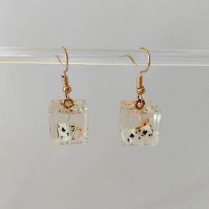 May include: A pair of clear resin cube earrings with gold-coloured ear wires. Each cube contains playing card suit symbols. The earrings are suspended against a neutral background.