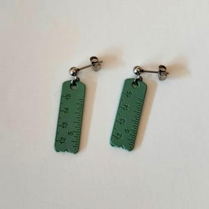 以下が含まれることがあります： 緑色の定規のイヤリングのペア。 各イヤリングは、5から15までの測定値を特徴とする定規のデザインの長方形の形状をしています。 イヤリングは、銀色のポストとボールスタッドが付いています。 白い背景にあります。