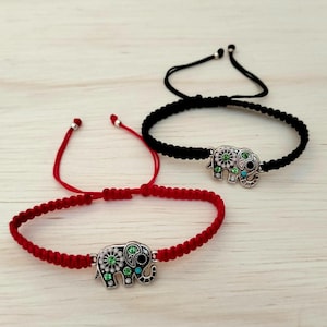 Puede incluir: Dos pulseras ajustables con abalorios de elefante. Una pulsera es roja, la otra negra. Cada abalorio de elefante plateado está decorado con piedras preciosas verdes y turquesas. Las pulseras se muestran sobre una superficie clara con vetas de madera.