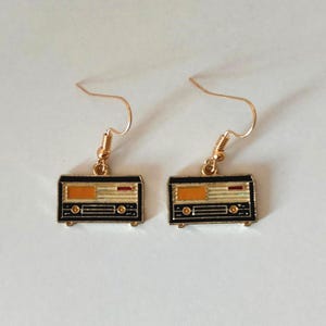 Puede incluir: Un par de pendientes dorados con forma de cinta de cassette. Cada pendiente presenta un diseño rectangular negro con detalles en naranja y rojo, y ganchos dorados.