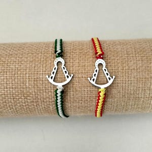 Könnte beinhalten: Zwei handgefertigte Armbänder mit einem silbernen Anhänger in Form einer religiösen Figur. Eines hat ein grün-weiß-schwarzes geflochtenes Band, das andere ein rot-gelb-weißes geflochtenes Band. Die Armbänder werden auf einer beigen Oberfläche präsentiert.