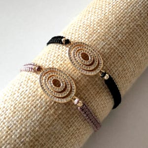 Puede incluir: Dos pulseras con colgantes ovalados dorados incrustados con pequeñas piedras transparentes. Los colgantes tienen un diseño en espiral. Una pulsera tiene un cordón negro y la otra un cordón malva. Se exhiben sobre una superficie beige texturizada.