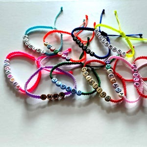 Puede incluir: Una colección de pulseras de amistad de colores con nombres escritos en cuentas negras. Las pulseras están hechas con cordones de diferentes colores y presentan nombres como "Victoria", "Nicolas", "Roberto", "Cayetana", "Paola", "Emma", "Alvaro", "Cristina", "Manuel", "Alejandro".