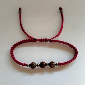 Op de afbeelding: Rode macramé armband met drie bruine, gespikkelde kralen en zilverkleurige accenten. De armband heeft een verstelbare schuifknoopsluiting en is gemaakt van een rood koord.