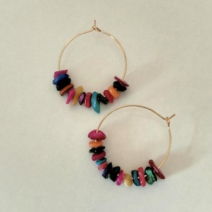 Op de afbeelding: Een paar goudkleurige oorringen met een reeks kleine, onregelmatige, veelkleurige stenen kralen die langs de onderkant van elke ring zijn geregen. De kralen hebben tinten roze, blauw, zwart, oranje en geel met een gepolijste afwerking.