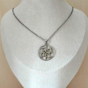 Puede incluir: Collar con colgante plateado con un colgante circular con un diseño geométrico. El colgante cuelga de una delicada cadena. El collar se exhibe sobre un busto blanco.