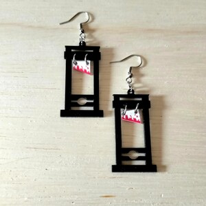 Puede incluir: Un par de pendientes de fantasía con un diseño de guillotina negra. Cada pendiente tiene un gancho plateado y una pequeña cuchilla rectangular con un patrón rojo y blanco. Los pendientes están colocados sobre un fondo claro de vetas de madera.