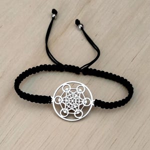 Puede incluir: Una pulsera trenzada negra con un colgante plateado del Cubo de Metatrón. La pulsera ajustable tiene un cierre de nudo corredizo. El colgante presenta un diseño geométrico detallado. La pulsera está sobre una superficie de madera clara.