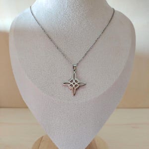 Op de afbeelding: Een zilverkleurige hangerketting met een vierpuntig sterontwerp met ingewikkelde details. De hanger hangt aan een fijne ketting, gepresenteerd op een witte buste. De ketting is een sieraad.