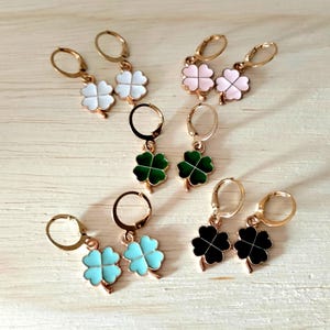 May include: Several pairs of gold-tone hoop earrings, each with a four-leaf clover pendant. The clovers are in white, pink, green, black, and light blue. The earrings are presented on a light-coloured wooden surface.