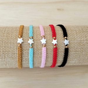 Può includere: Sei braccialetti dell'amicizia colorati. Ogni braccialetto presenta un cordoncino intrecciato in diversi colori: beige, azzurro, rosa, rosso e nero. Ogni braccialetto ha un ciondolo a forma di stella bianca e perline dorate. I braccialetti sono esposti su un tessuto beige.