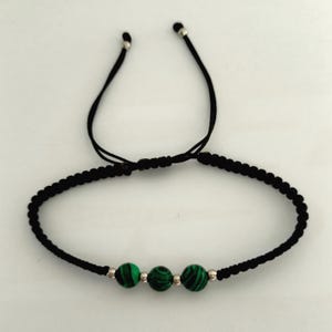 May include: A black adjustable bracelet featuring three green and black malachite beads. The bracelet has a braided design with silver-colored metal accents. The adjustable cords are black.