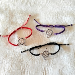 Puede incluir: Tres pulseras ajustables con un dije de nudo celta plateado. Las pulseras son rojas, moradas y negras.