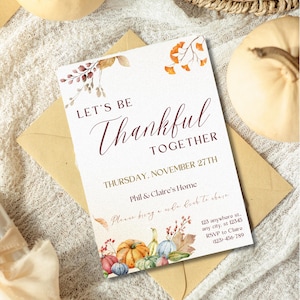 Könnte beinhalten: Danksagungseinladung mit dem Text „Lasst uns dankbar sein“ und floralen und Kürbisillustrationen. Die Einladung befindet sich auf einem cremefarbenen Hintergrund mit einem braunen Umschlag, umgeben von Kürbissen und einem Weidenkorb.