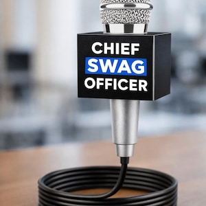 May include: A silver microphone with a black rectangular box that reads "CHIEF SWAG OFFICER" in white text on a black background with a blue stripe. The microphone cable is coiled on a wooden surface.