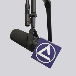 May include: A black microphone with a foam windscreen is suspended from a black boom arm. A square, blue sign with a white triangle inside a white circle is attached to the microphone.