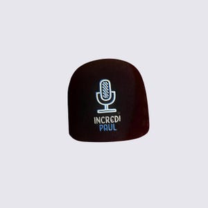 Puede incluir: Una funda negra para micrófono con un gráfico de micrófono blanco y azul claro y el texto "INCREDI PAUL". La funda está hecha de un material suave similar a la espuma.