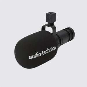 Puede incluir: Un micrófono Audio-Technica negro con un filtro de espuma. El micrófono tiene un cuerpo cilíndrico y una pieza de montaje cuadrada. El nombre de la marca está impreso en blanco en el filtro.