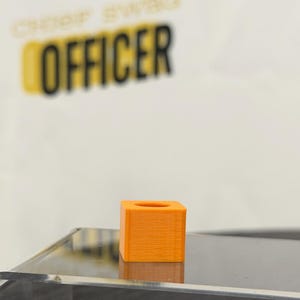 May include: An orange, cube-shaped object with a circular hole on top. The object is sitting on a reflective surface. The text "CHEER SWAG OFFICER" is blurred in the background.