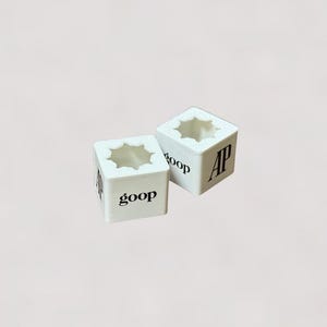 May include: Two white, cube-shaped objects with a star-shaped opening on top. The word "goop" is printed in black on one side of one cube, and "AP" is printed on the other. The cubes are on a light pink background.