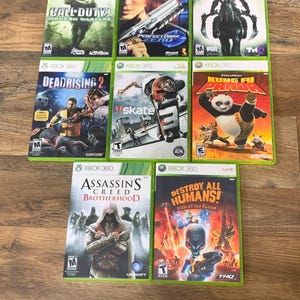 May include: A collection of eight Xbox 360 video game cases. Titles include "Call of Duty 4: Modern Warfare," "Perfect Dark Zero," "Darksiders II," "Dead Rising 2," "Skate," "Kung Fu Panda," "Assassin's Creed Brotherhood," and "Destroy All Humans!"