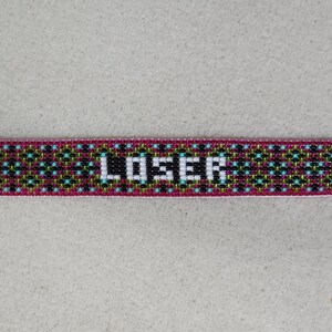 May include: A beaded bracelet with the word "LOSER" in white block letters. The bracelet has a pink border and a geometric pattern in black, teal, and gold. The bracelet is rectangular in shape.