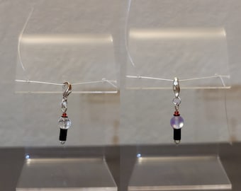 Puede incluir: Un charm plateado con un cierre de mosquetón, con una cuenta transparente, una cuenta cilíndrica negra, una cuenta roja y una cuenta morada. El charm se exhibe en un soporte de acrílico transparente.