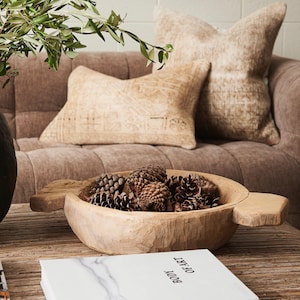 May include: A wooden bowl filled with pine cones sits on a wooden table. Two decorative pillows rest on a brown sofa in the background. A book with the text "BODY OF ART" is in the foreground.