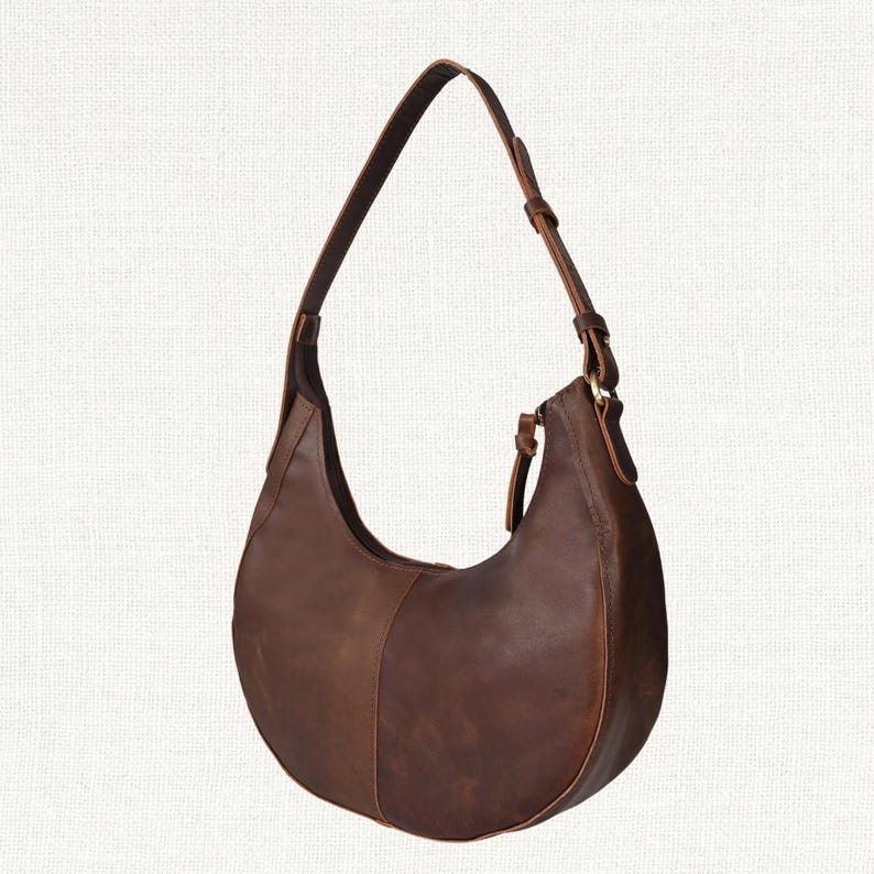 Puede incluir: Un bolso de hombro en forma de media luna de cuero marr&oacute;n con una correa larga. El bolso tiene un dise&ntilde;o curvo con cierre de cremallera y un panel contrastante de cuero marr&oacute;n m&aacute;s oscuro. El bolso est&aacute; dise&ntilde;ado para llevarse al hombro.