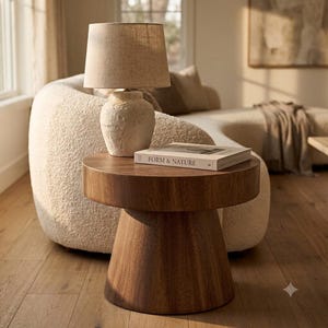 May include: A round wooden side table with a pedestal base, topped with a textured off-white ceramic lamp featuring a beige fabric shade and a hardcover book titled FORM & NATURE resting on the surface of the table.