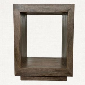 May include: A square, open-frame wooden shelf in a warm brown tone. The shelf has a rectangular base and a thick frame, creating a box-like structure. The wood grain is visible, adding texture to the piece. The design is simple and modern.